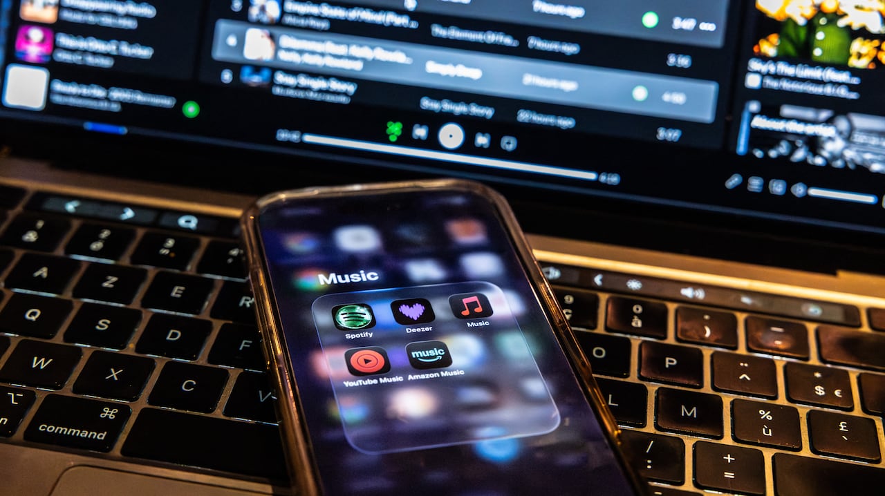 A phone shows music apps including Spotify and Deezer as it rests on the keyboard of a laptop, which appears to have a music program open on its screen.
