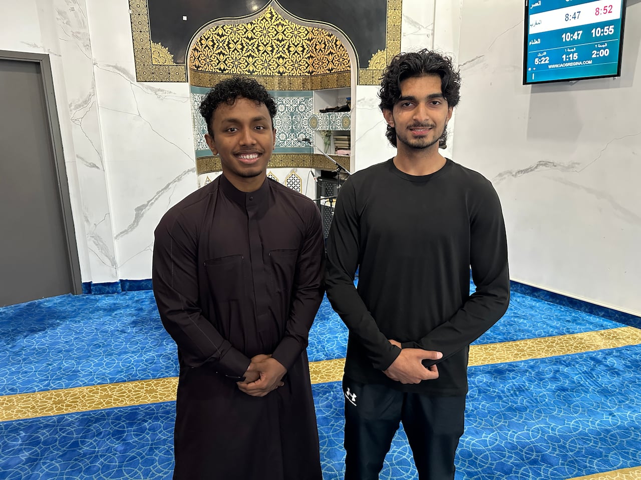 Two men stand next to each other for a photo inside the prayer hall of a mosque.