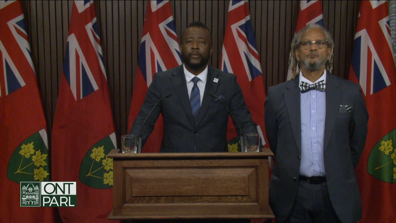 A man in a suit stands with two arms on a podium, while another man with glasses stands beside him.