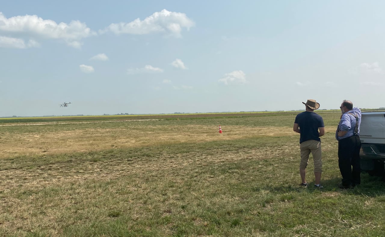 Two men stand near the back of a pickup truck in a field, watching a drone flying in the distance.