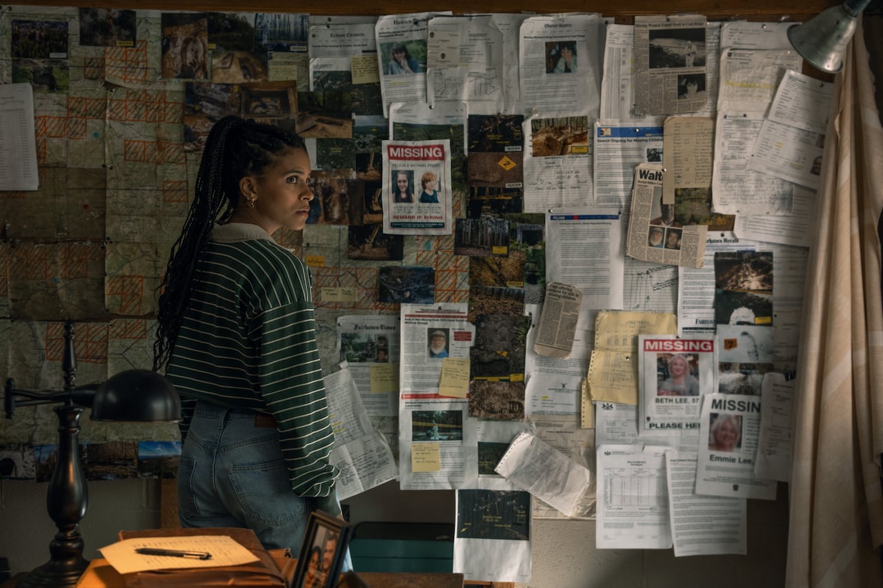 A woman stands in front of a corkboard with pictures and documents pinned to it.