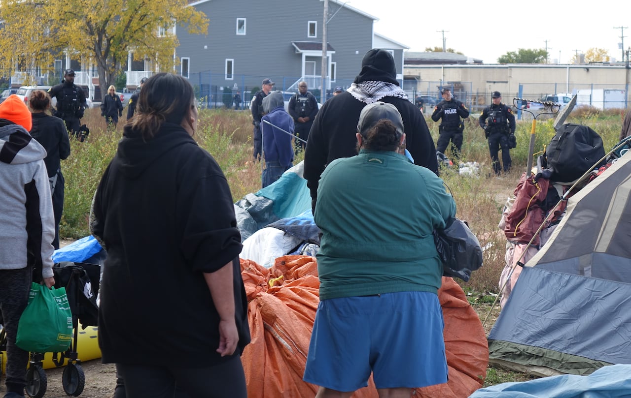People stand with their backs to the camera next to tents and piles of belongings as a line of police in black uniforms move toward them.