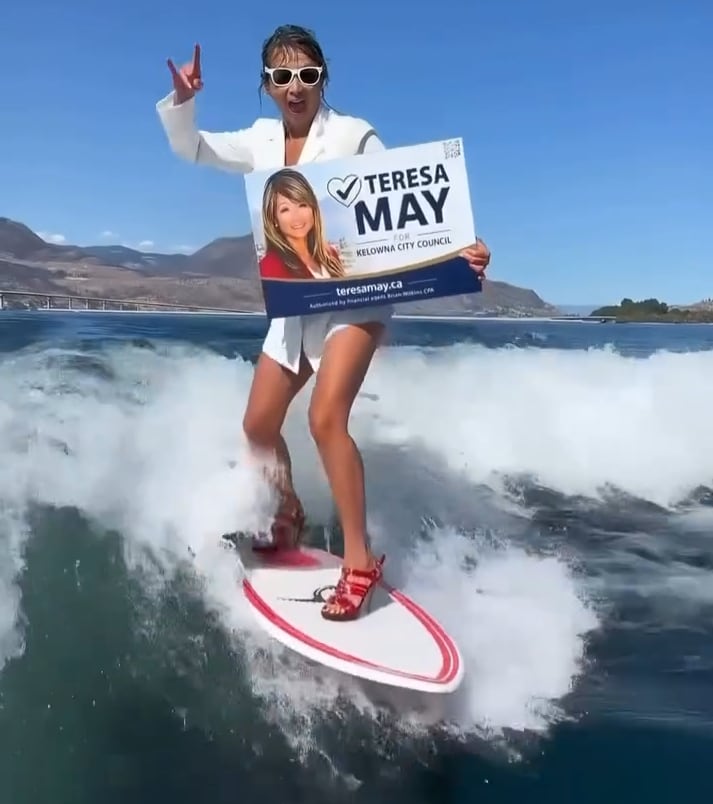 A woman in a blazer and red high heels stands on a wake surf board holding a campaign sign that says Teresa May