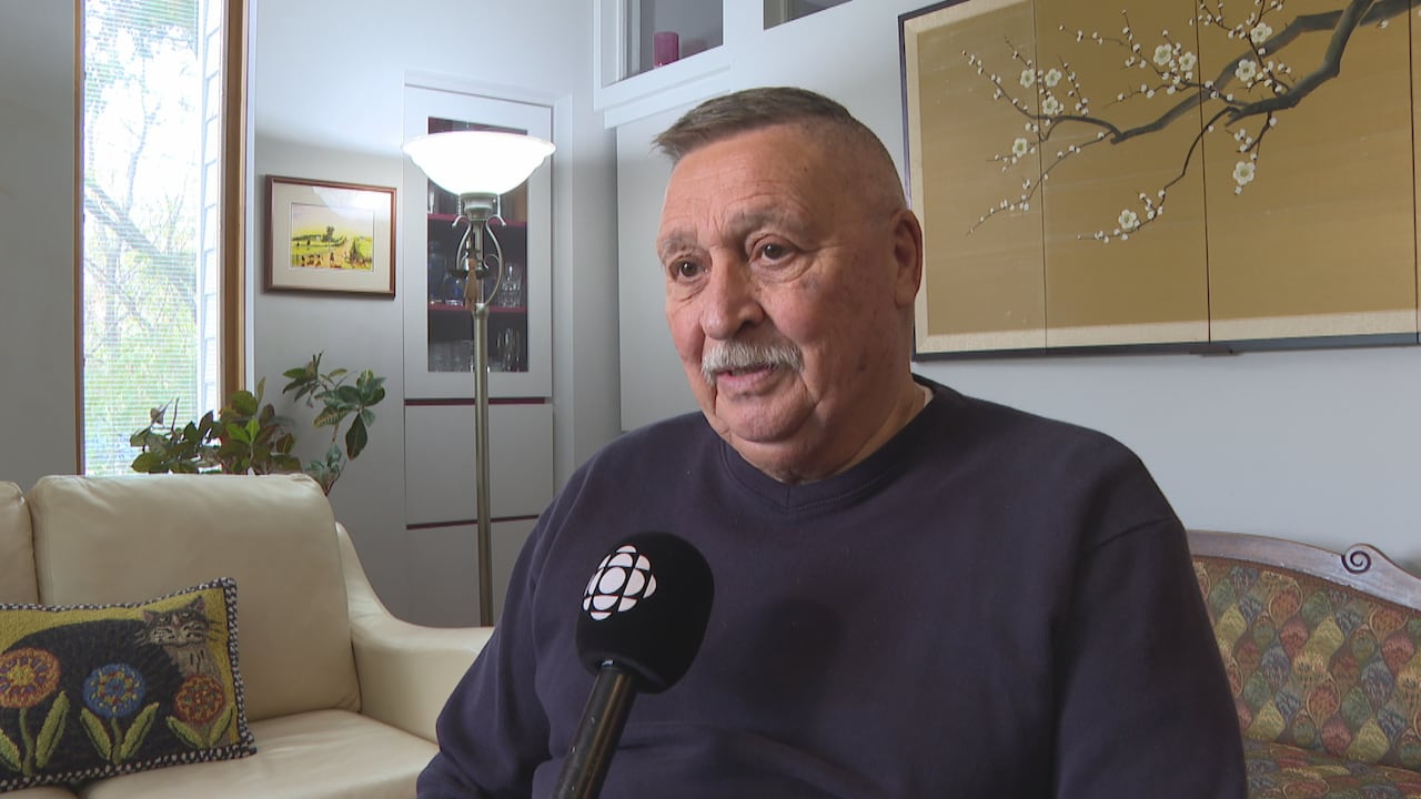 A man in a dark shirt with short hair and a moustache looks off camera. He's sitting indoors talking into a microphone.