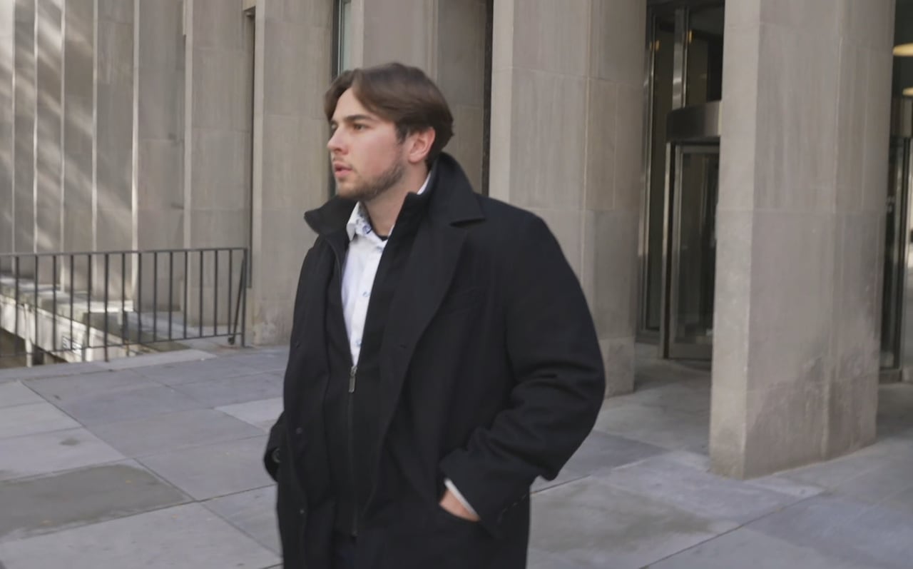 Man with brown hair and black jacket looks away from the camera outside a concrete building.