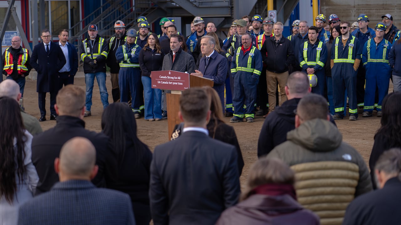 A man at a podium speaks to a crowd while workers are in the background.