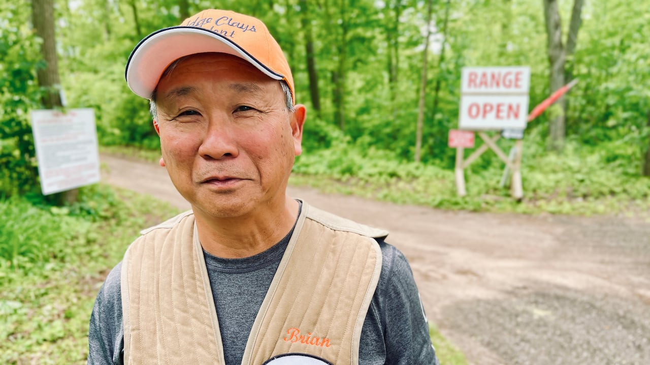 Brian Yee wearing a beige vest and orange hat. He is in a wooded setting, with a "range open" sign visible behind him.