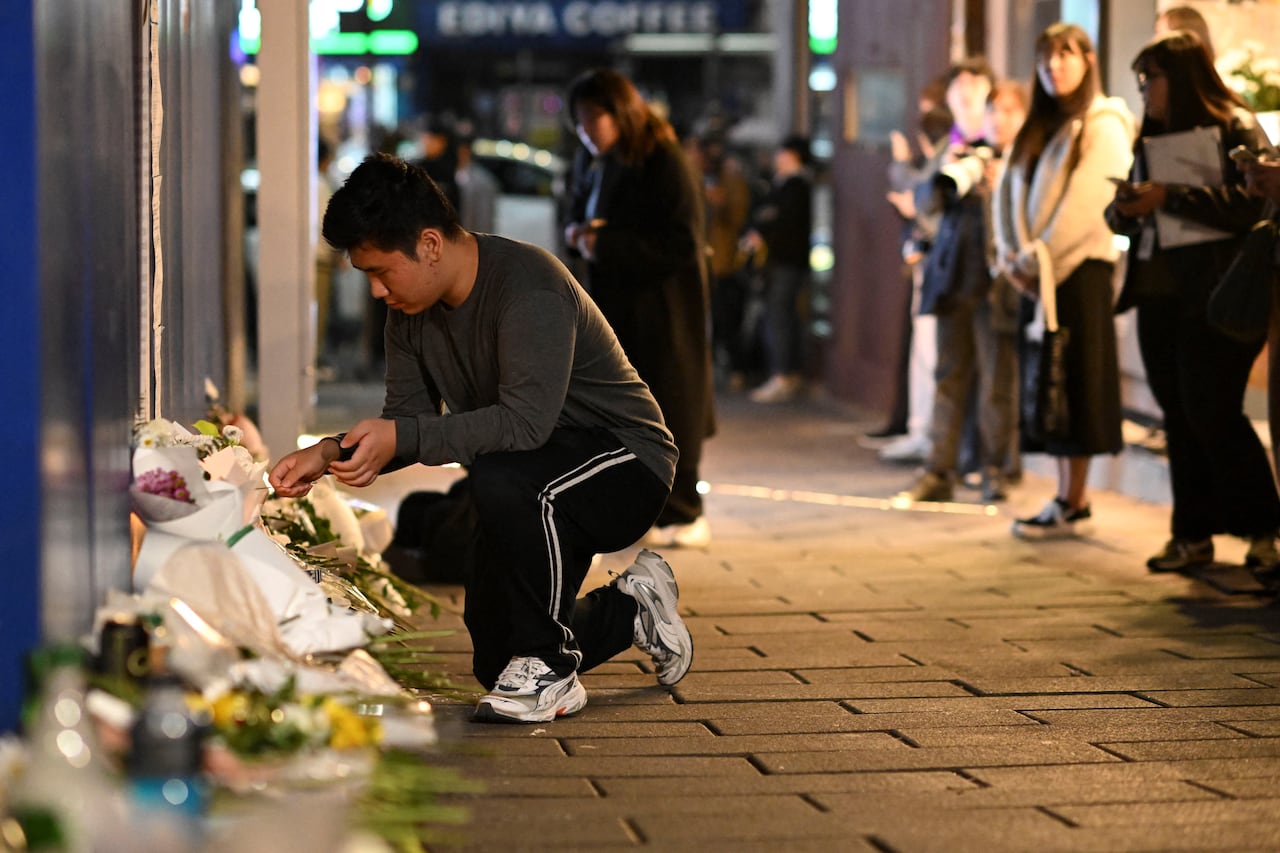 A young man kneels next to a pile of flower bouquets laid along the side of a building on a stone alleyway.