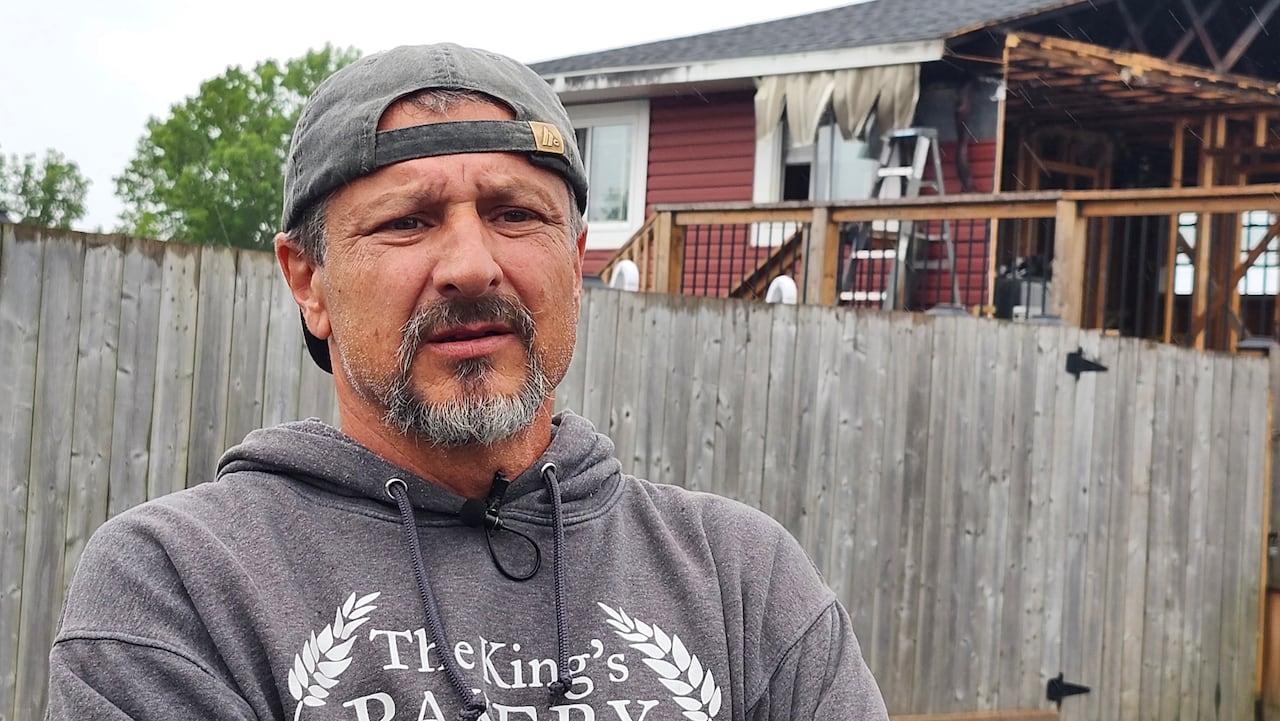 Man stands with arms crossed in front of burned out home.