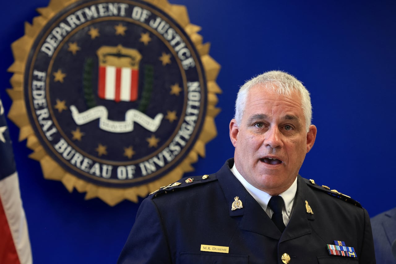 A man with white hair in a police uniform stands at a wood lectern in front of a blue background.