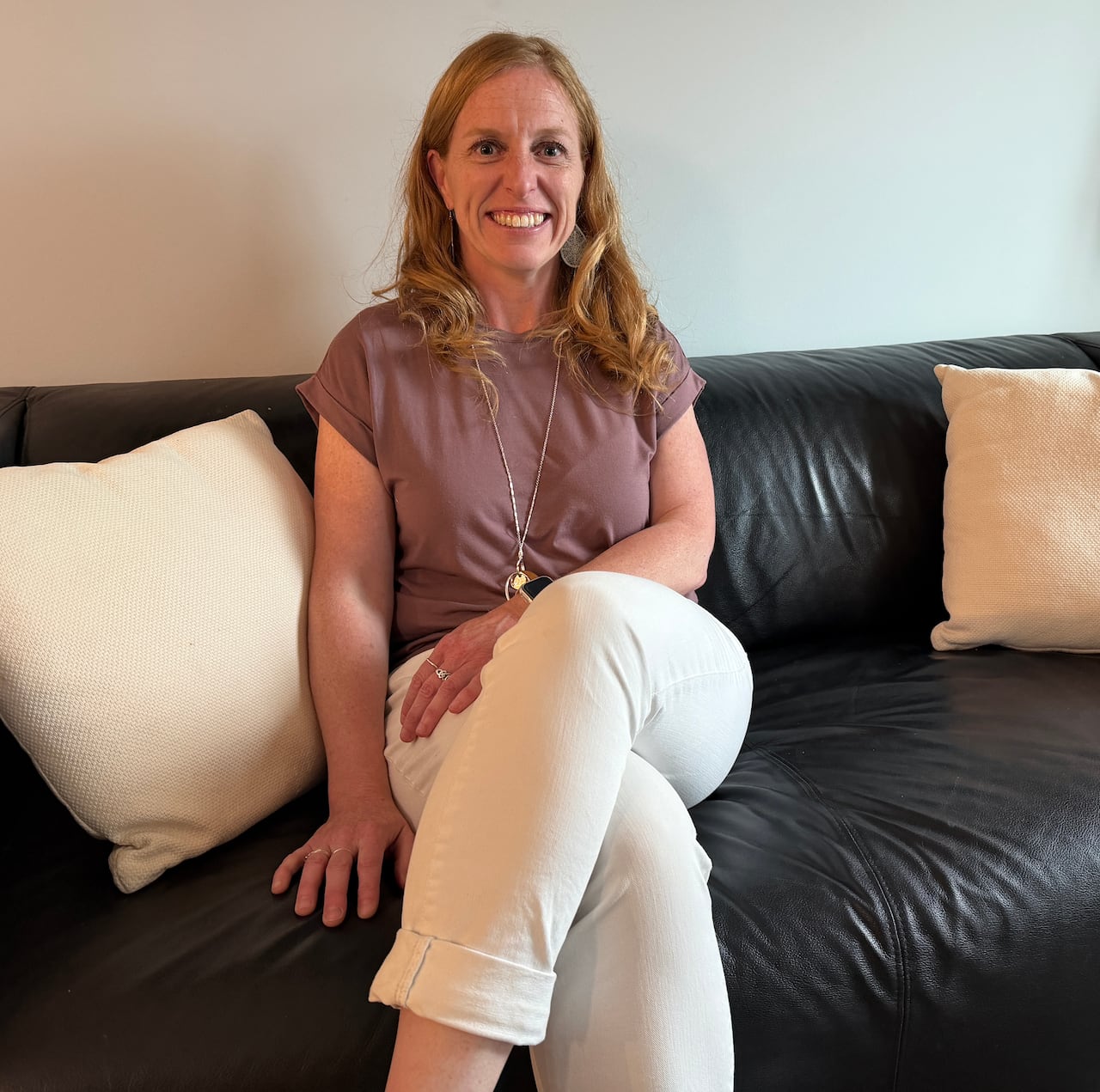 Woman smiling sitting on a sofa wearing white jeans and a mauve t-shirt.