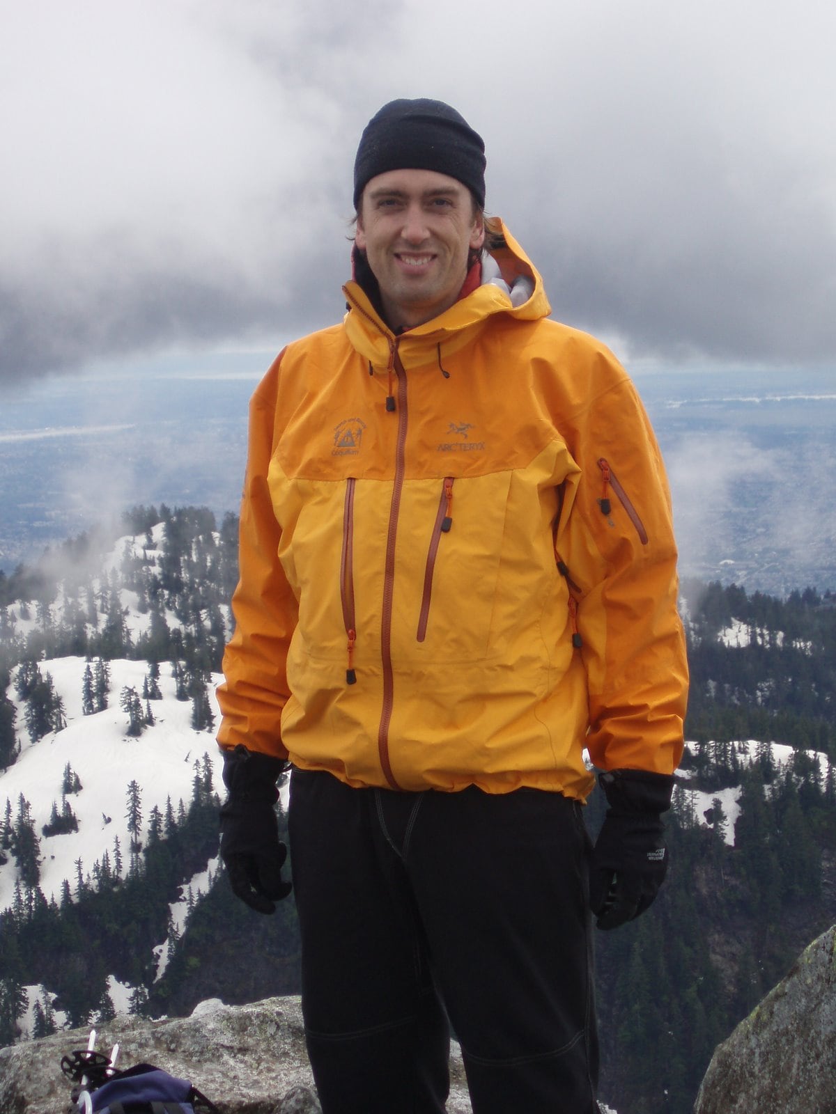 A man in a black toque and orange jacket is pictured standing on what appears to be a mountaintop, a snowy tree-topped landscape behind him.