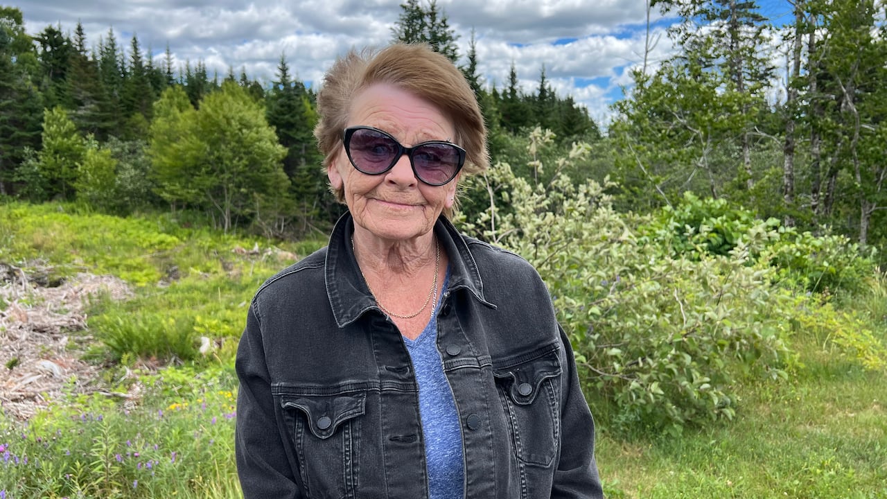 A woman in sunglasses, with red hair, wears a jean jacket with a blue shirt. She stands outside in front of greenery.