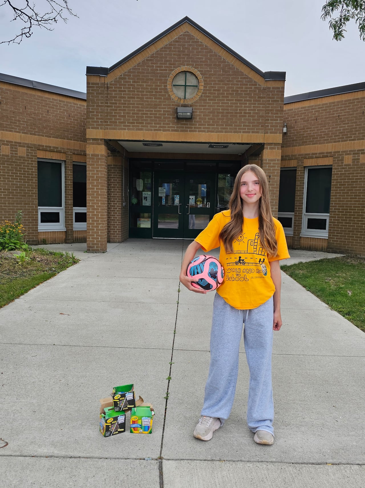 A young girl stands in front of a brick building. She is holding a soccer ball in her hand. On the ground next to her are several boxes of crayons.