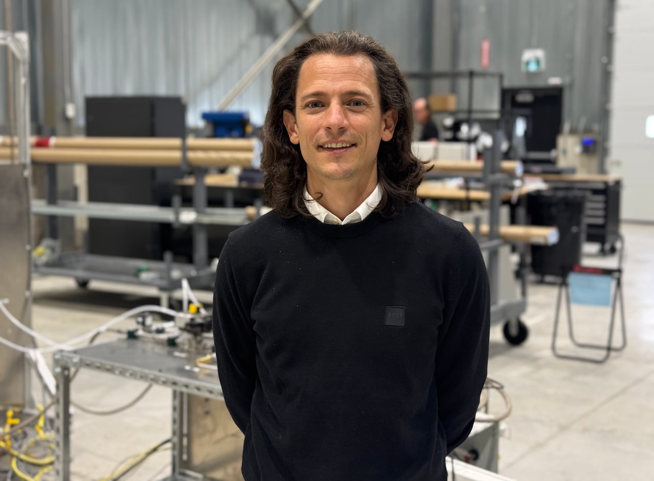 a man with long hair and a collared shirt sticking up from under a black sweater smiles for the camera in a workshop type building