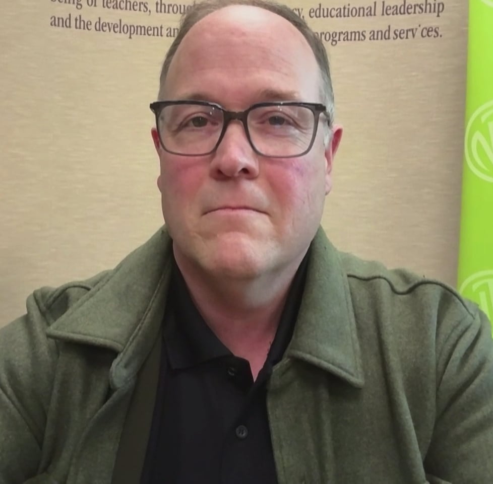 A man with glasses sits with the backdrop of N.L. Teachers' Association background.