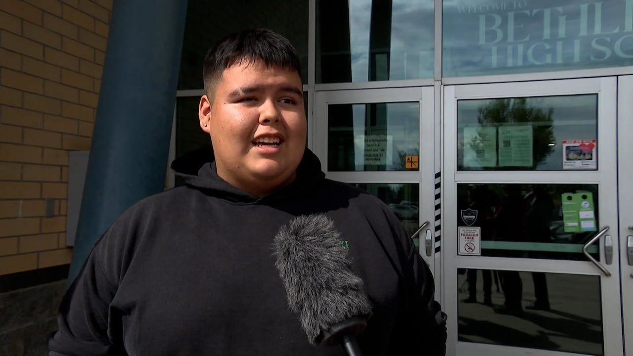 A young man with black hair in a black hoodie standing outside the doors of a high school.