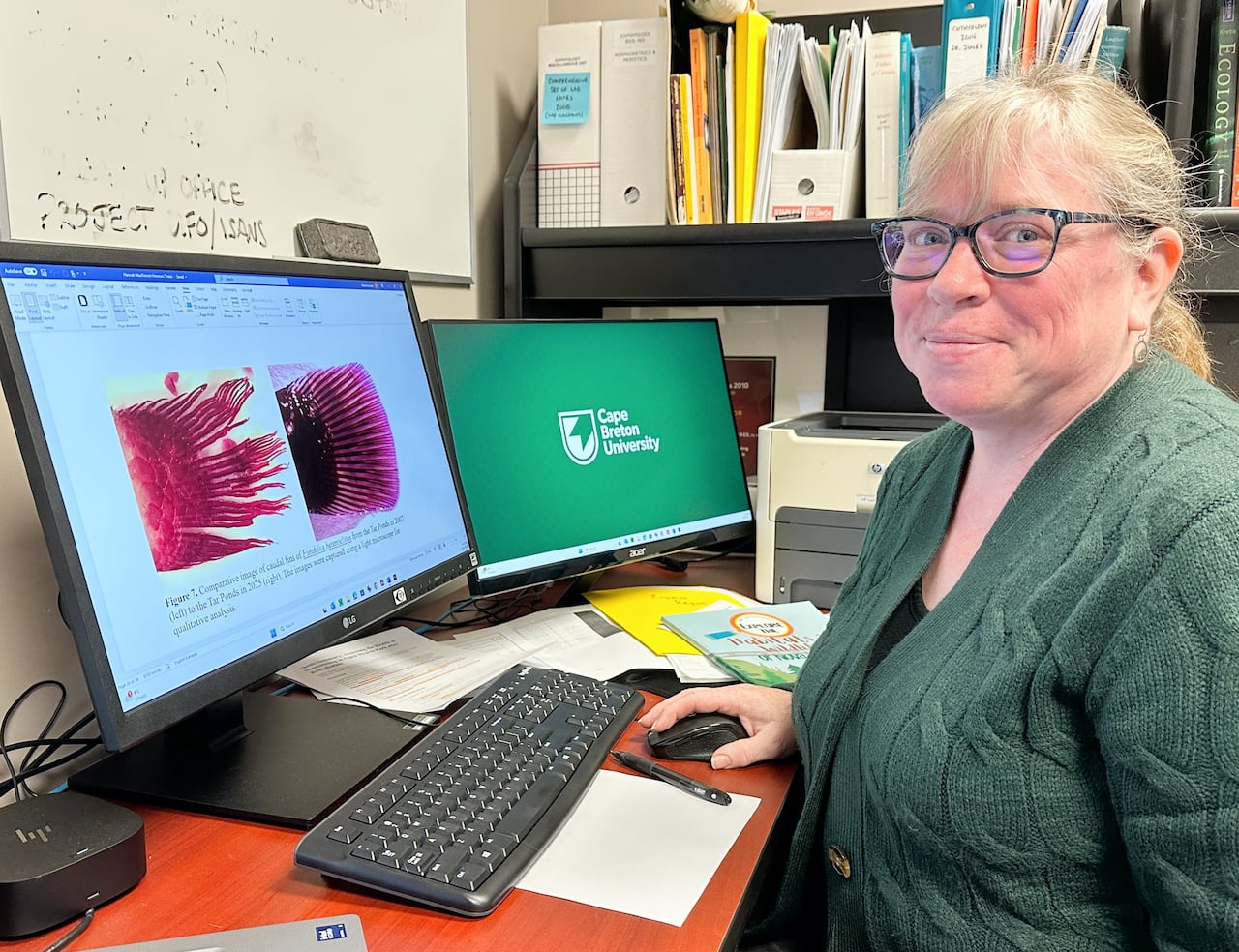 A woman in a green sweater wearing glasses sits in an office at a computer with two screens. One says Cape Breton University and the other shows two fish fins.