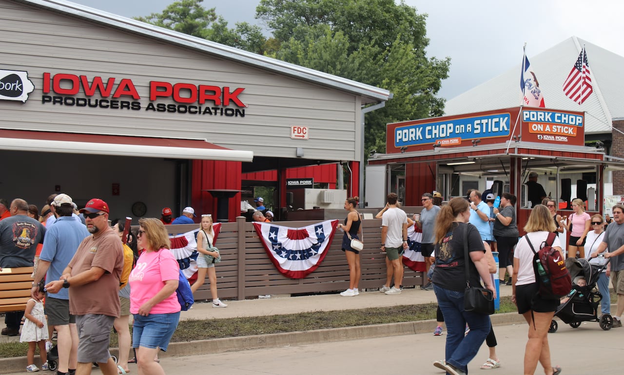 A crowd of people walk around buildings on a fairground. One building has a sign that reads "Iowa Pork Producers Association" and there is a trailer that reads "Pork Chop on a Stick"