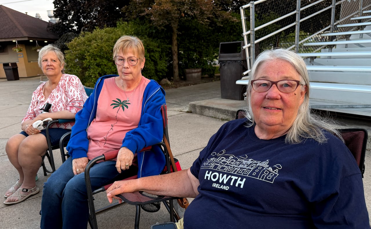 Three women sit in folding chairs in a parking lot.