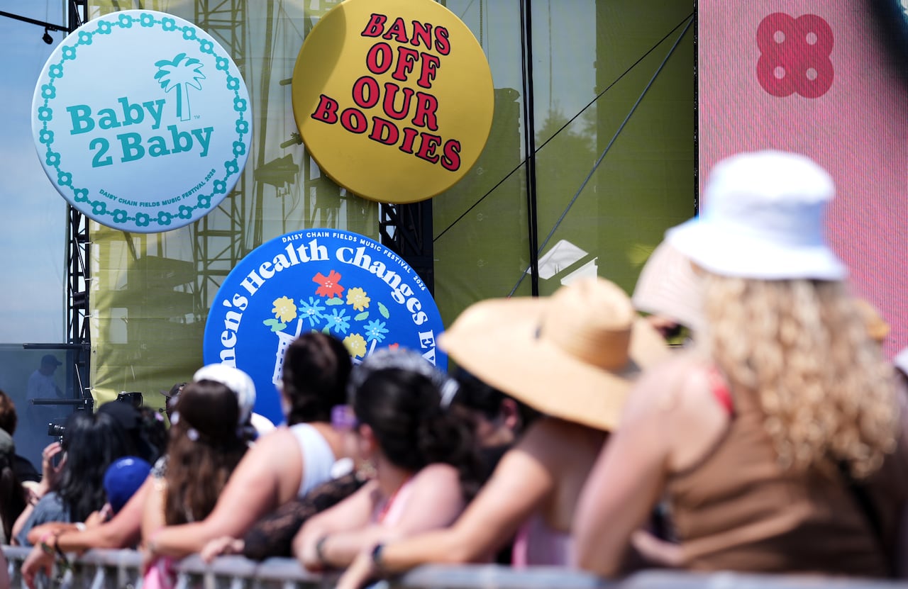 A line of people are shown at the barrier at a concert, with three large circle displays visible beyond them, one saying "Bans off our bodies," another saying "Baby 2 Baby".