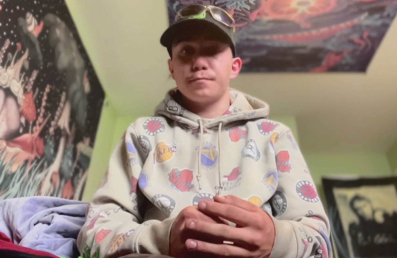 A young man wearing a hat, sits on a bed inside a small room.