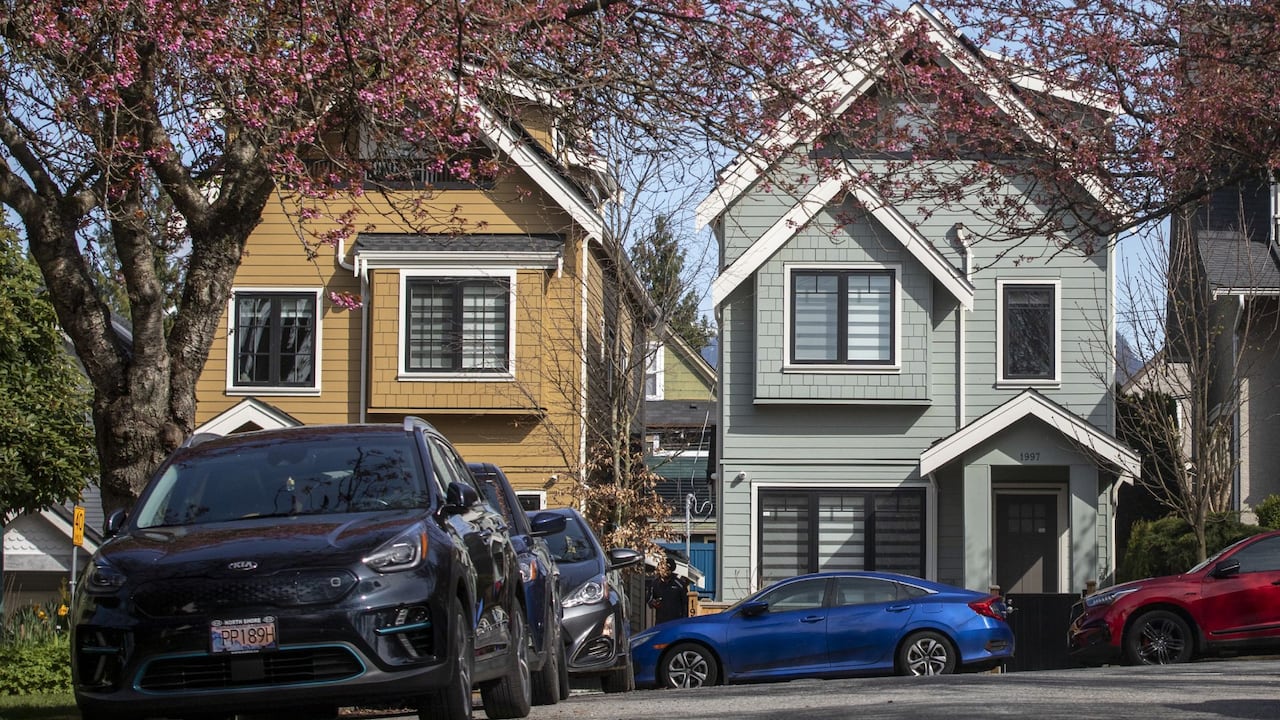 Two houses are pictured. The one on the left has yellow wood panelling. The one on the right is grey. A black Kia car is parked in the foreground.