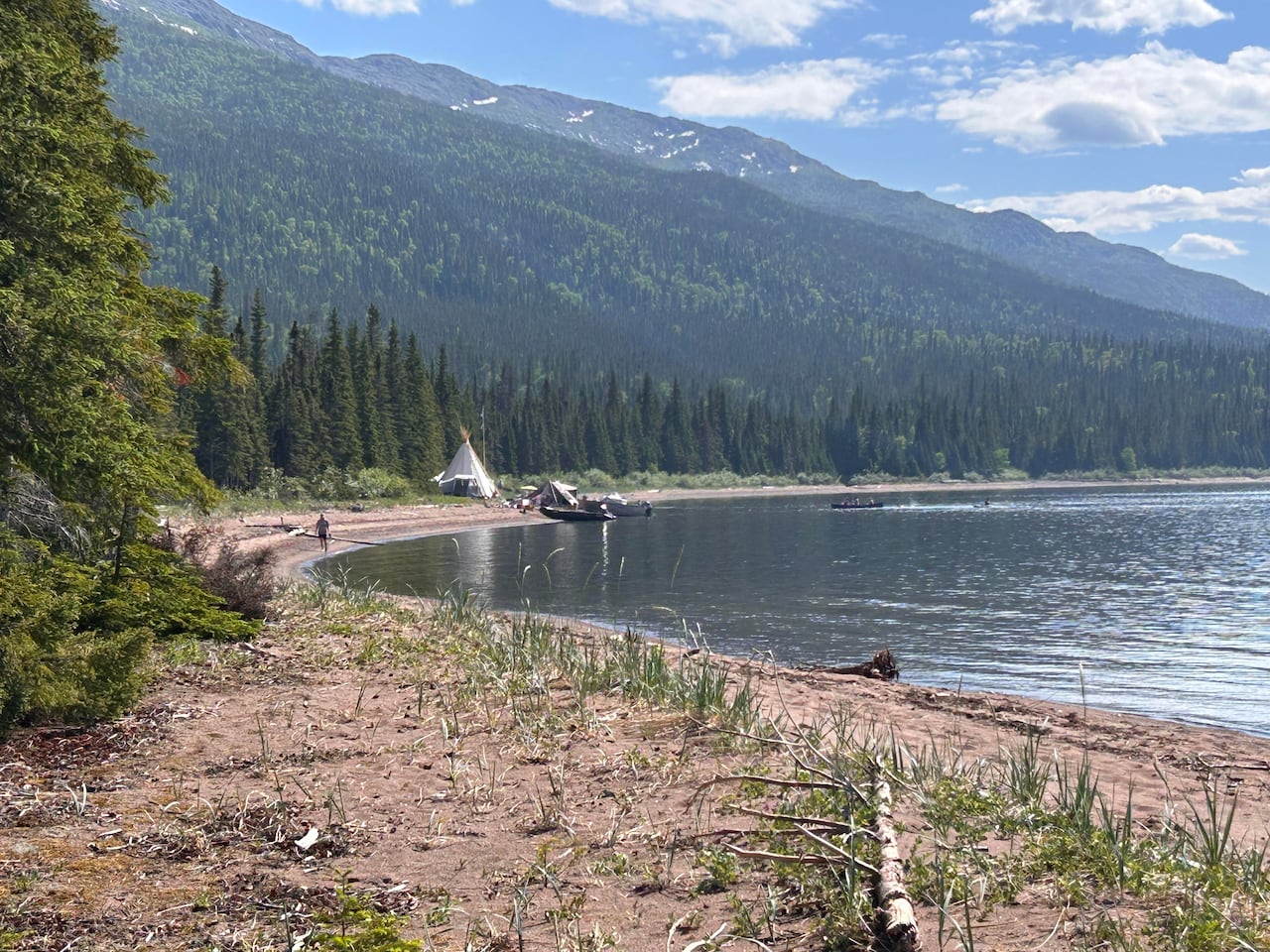A traditional Innu tent is pictured on a sandy beach, on a sunny day in a mountain range.
