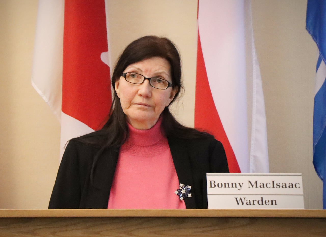 A woman sits at a desk next to a nameplate that says "Bonny MacIsaac, Warden" with flags behind her.