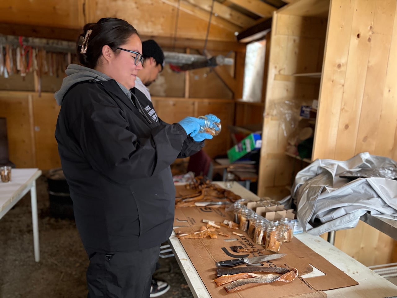 Kwägąya, Azzy Allen, wearing glasses and blue gloves, holds a glass jar while standing beside a table covered with smoked fish.