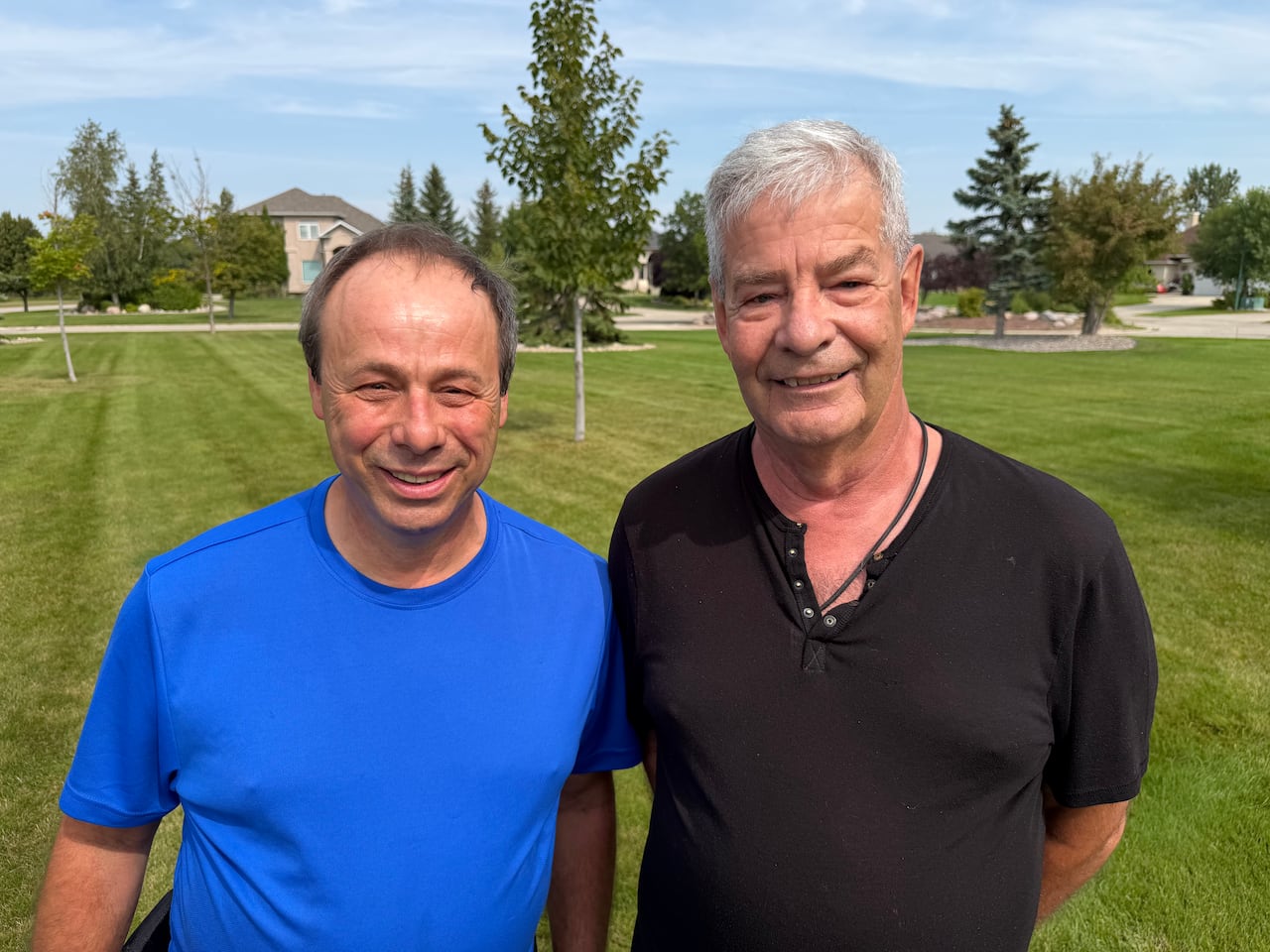 A man wearing a blue t-shirt stands next to a man with a black t-shirt on a lawn in a residential area
