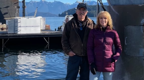A man and a woman stand together on a dock at the edge of water, with mountains in the background.