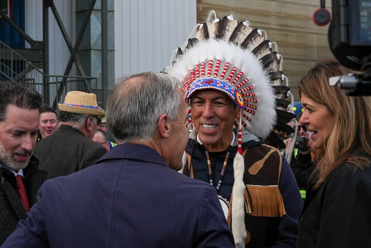 A man and a woman speak to a man in an Indigenous headdress.