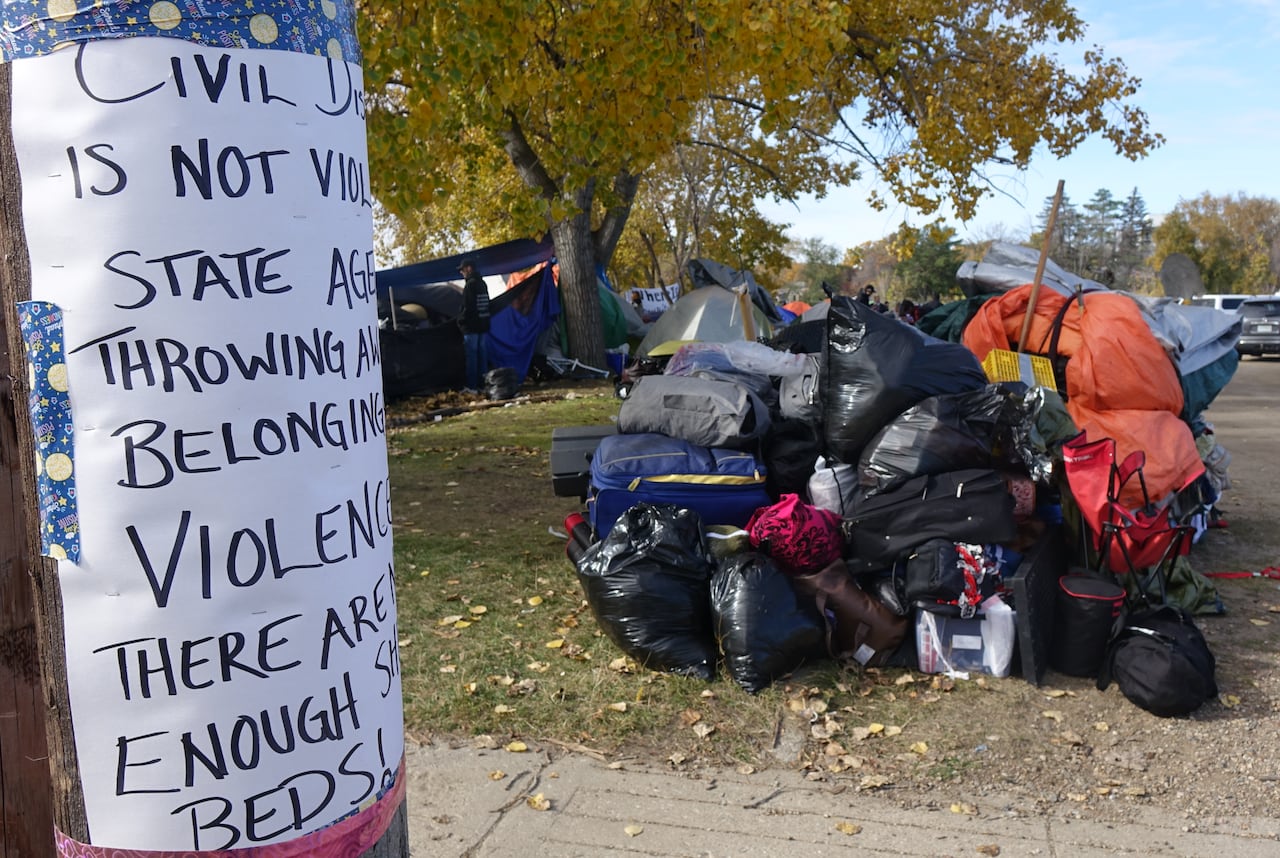 A handwritten protest sign taped to a power pole in the foreground, with a pile of camping gear and belongings next to it.
