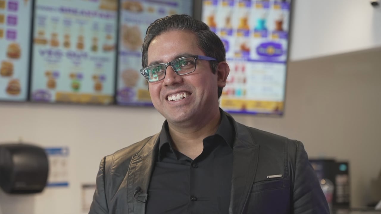a man with glasses and a grey suit smiles at someone off camera while standing in a fastfood restaurant, the background blurred