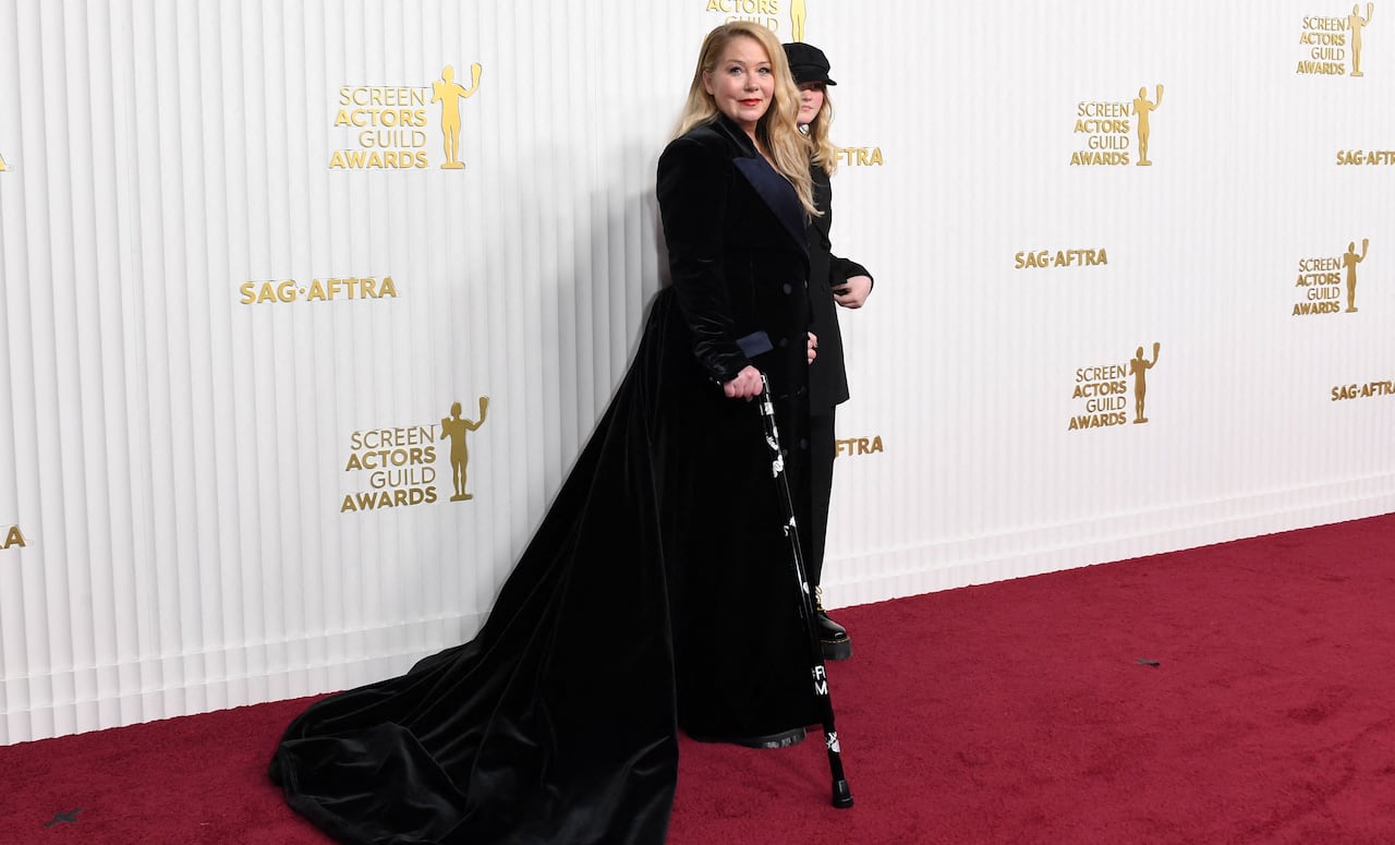A woman in a large black dress and carrying a black cane, poses on a red carpet.