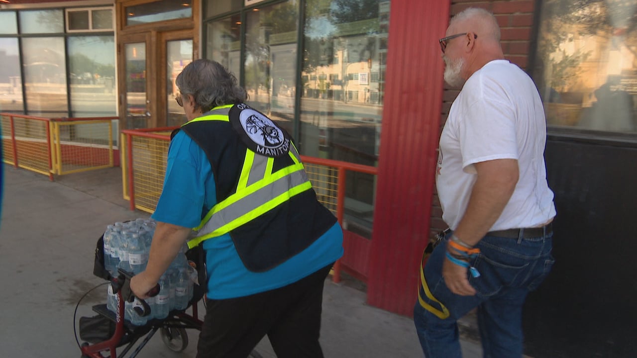 A woman with a walker filled with cases of water bottles walks along a city street with a man in a white t-shirt