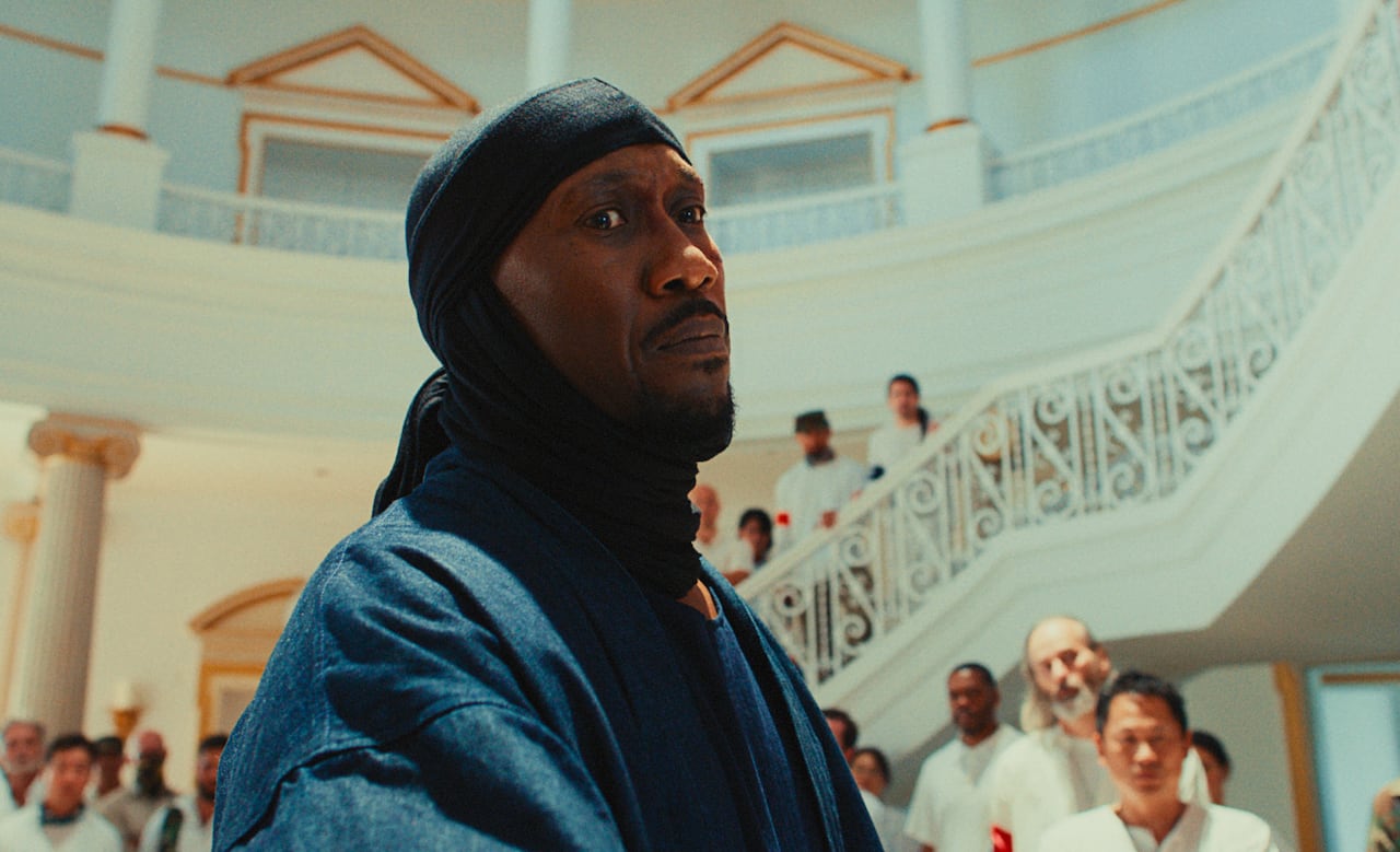 A man stares intently toward the camera. Behind him is a white winding staircase, on which people dressed all in white stand looking at him.