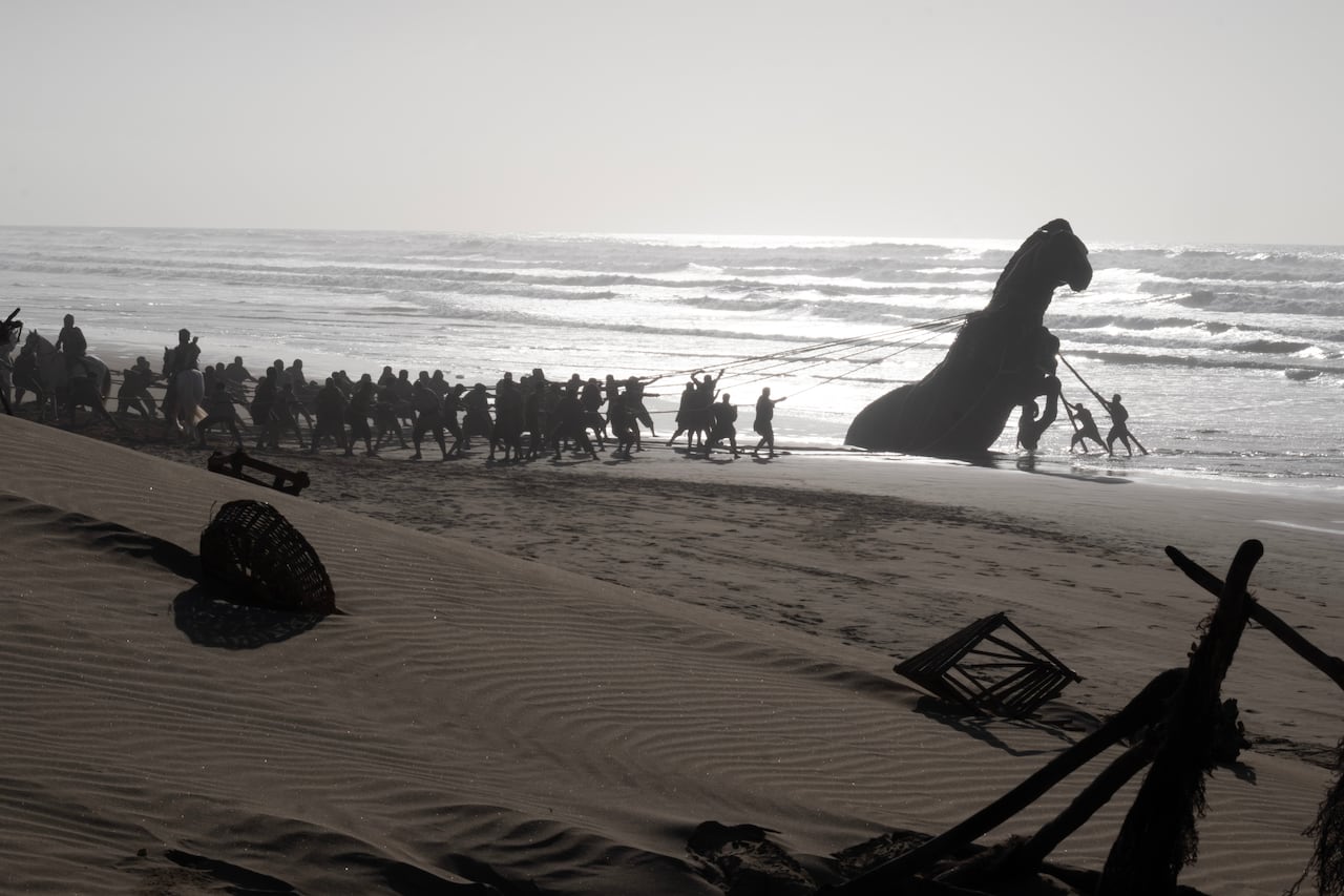 hundreds of men labour to pull a large statue of a horse from the sand into a palace.