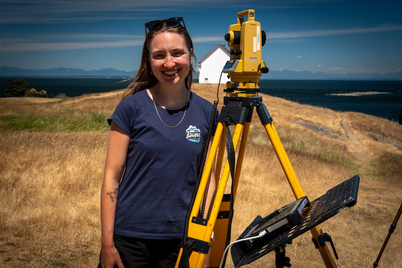 Lauren is a 20 something year old white female with long dirty blonde hair. She is standing on an island and you can see the Salish Sea behind  her. Next to her is a yellow piece of surveying equipment.
