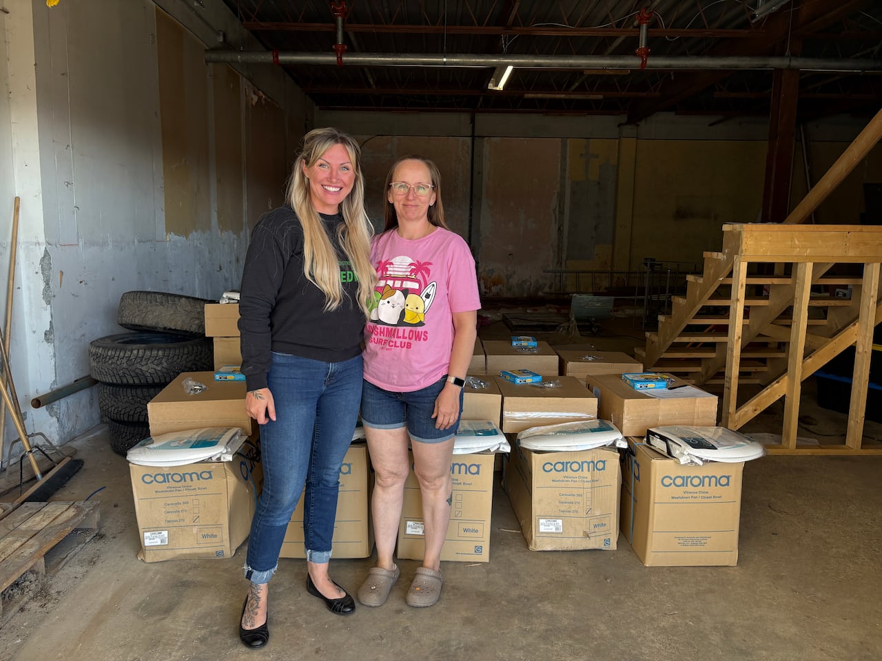 Two women stand together in front of numerous card board boxes and packaged toilet seats.