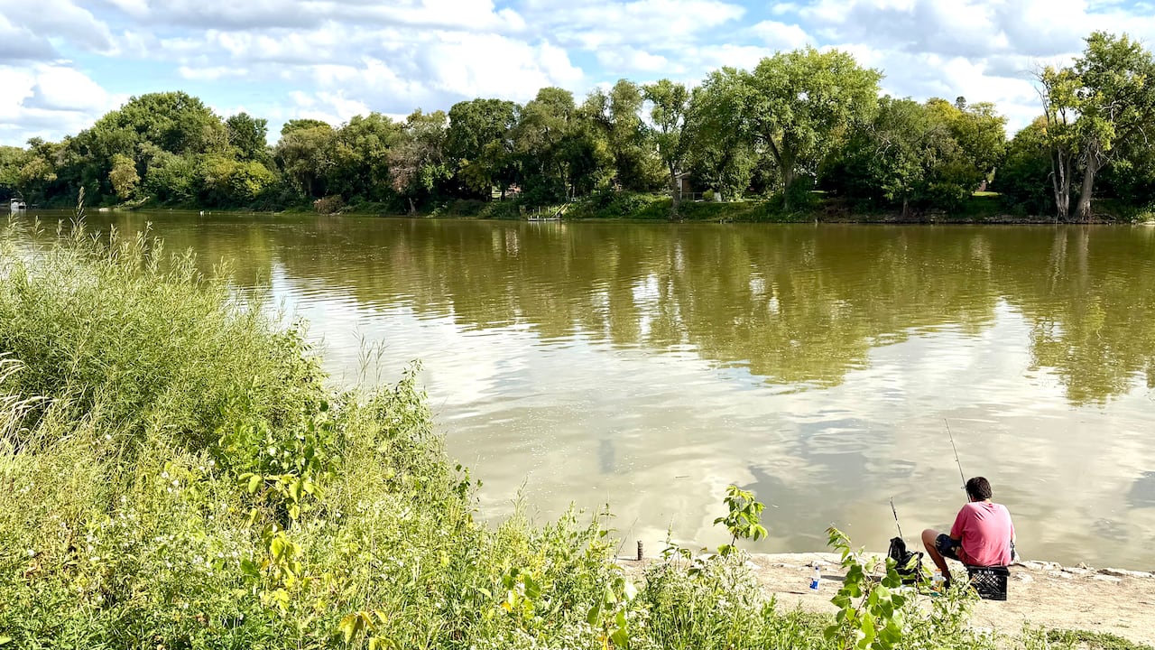 A person fishes on the tree-lined bank of a river.