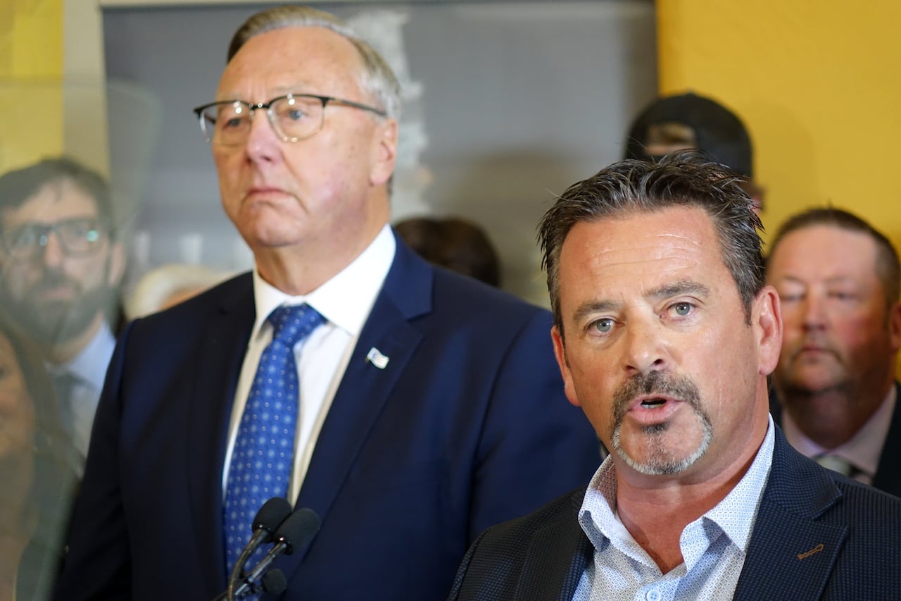 A man wearing a suit speaks into a microphone. A taller man in a suit is standing to his right.