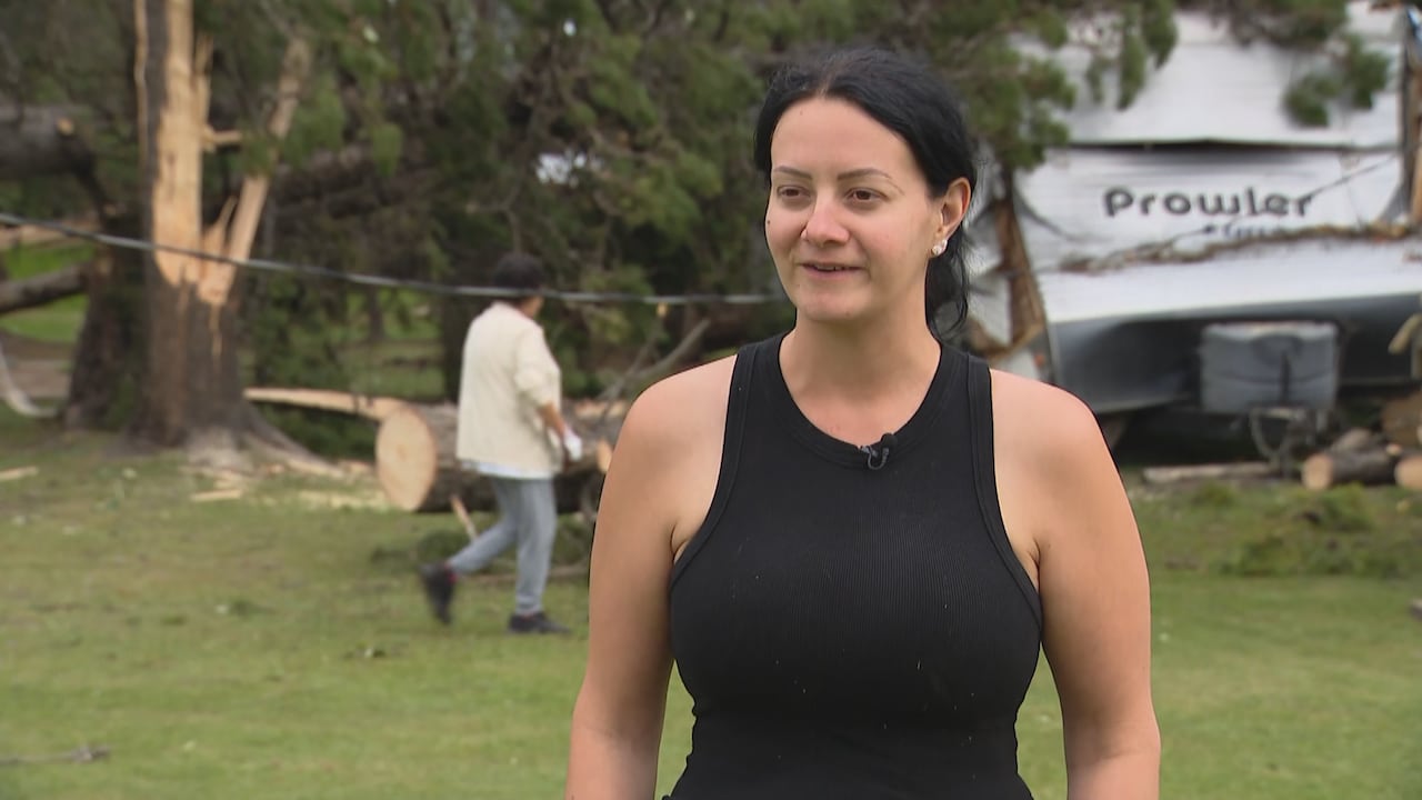 A woman with black hair, wearing a black tank top, looks at a camera. Behind her are many fallen trees and a person, wearing gloves, preparing to clean up.