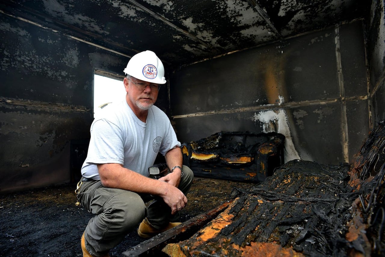 A man wearing a white hard hat and t-shirt crouches in a room heavily damaged by fire near burnt furniture