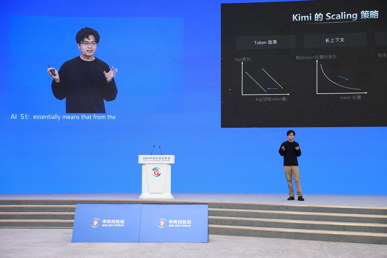 a man with glasses and khaki pants speaks on stage in front of a screen with english and chinese characters describing an AI function