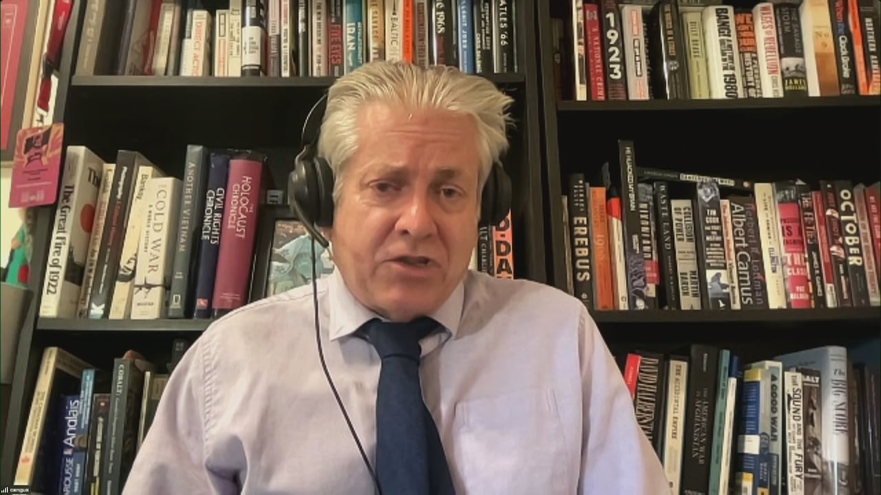 Man with white hair, wearing headphones, a white shirt, and a dark blue tie, zooms from a room with tall bookshelves.