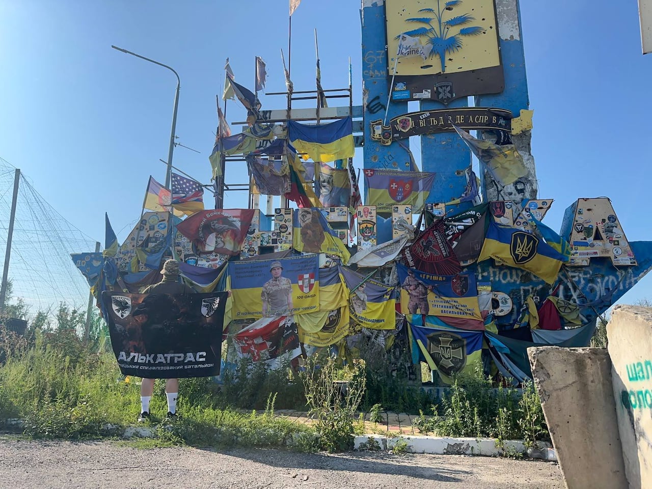 Person holding up the banner of the Alcatraz Battalion in front of the iconic Donetsk Oblast entry stele