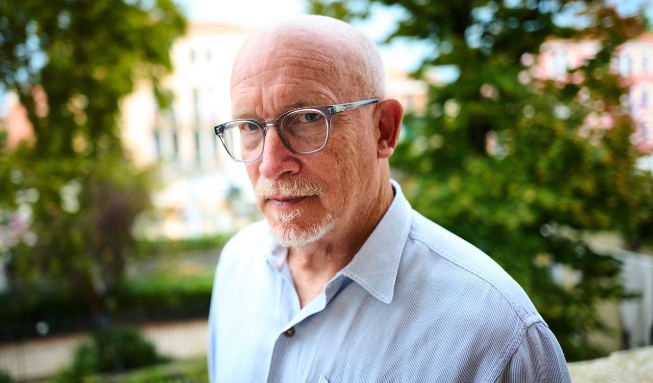 A bald man with glasses and a white goatee looks into the camera in a shoulder-up photo.