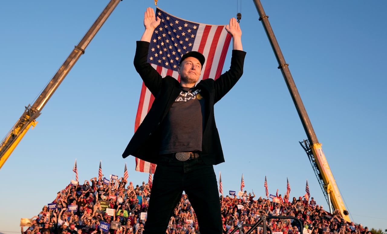 A man stands with his hands up and an American flag and a crowd on bleachers visible behind him.